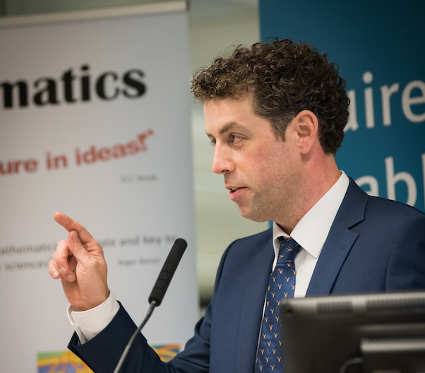 Photograph of a man wearing a navy suit gesturing while delivering a talk.