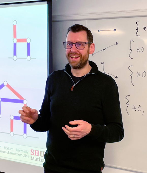 A man in a black jumper and glasses stands smiling next to a whiteboard and projector screen. The whiteboard has hand-drawn mathematical objects and the projector pre-rendered diagrams.